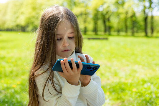 Little girl using or playing with a smartphone in nature park instead of walking and enjoying sunny day. Child watching videos on a smartphone device. Copy space