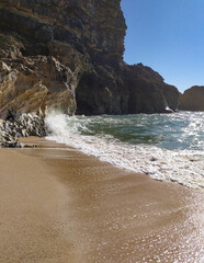 Waves crashing against rocky cliffs under a clear blue sky near the shoreline during midday sun