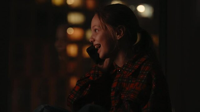Portrait of happy excited 10 yo girl with long hair in ponytail wearing red and black plaid shirt, smiling talking on smartphone sitting by window with city bokeh lights glowing outside, close-up.