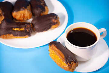 Chocolate-covered eclairs on a plate with cream filling sit next to a white cup of black coffee. One on a saucer, showing off its creamy cream.
