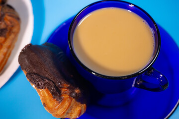 A blue glass mug filled with creamy coffee sits on a matching saucer, topped with chocolate-covered eclairs. A plate of more eclairs can be seen in the background. A sweet and comforting treat.