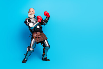 Elderly man dressed as a knight with boxing gloves posing against a bright blue background in a medieval costume and modern additions