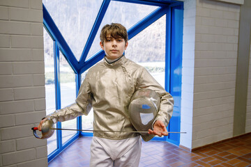 Teenager boy in fencing uniform with epee and mask. Teen fencer indoors, before competition...