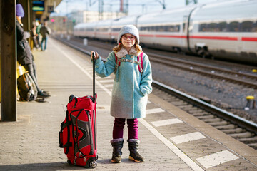 Little happy girl with glasses and a winter outfit standing on a train platform, holding a red...