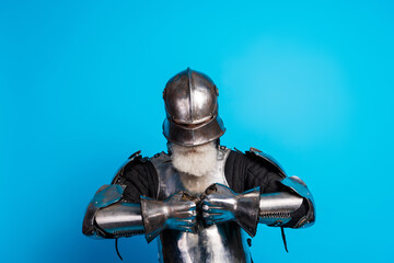 A senior man dressed in medieval armor adjusts his gauntlet against a vibrant blue backdrop,...