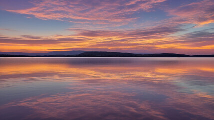 Obraz premium A serene sunset over a calm lake. Wispy clouds are reflected on the water's surface, creating a mirrored effect of the sky.