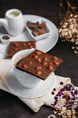 Coffee in a white cup with chocolate, flowers on a dark background, flatlay, chocolate with whole hazelnuts, handmade candies, coffee with milk, exquisite dessert