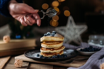 woman sprinkles blueberry pancakes with powdered sugar, breakfast, berries, vegan, dark background, healthy breakfast, pancakes with berries on fruit