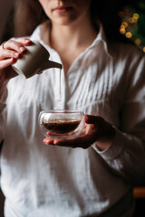 a girl in a white shirt pours milk from a white milk jug into an espresso coffee in a transparent cup, drops of milk in the air, bokeh, coffee with milk, a woman drinks coffee