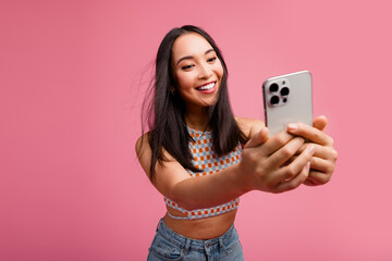 Happy young woman taking selfie with smartphone against a vibrant pink background, showcasing...
