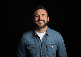 Obraz premium Studio portrait of a happy man wearing a denim shirt over a white t-shirt, set against a black background. He has short hair, a beard, and brown eyes, smiling with his teeth showing while looking dire