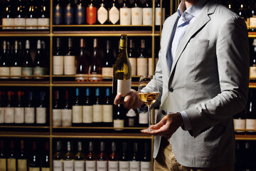 Man holds filled glass with white wine and opened bottle, standing against wooden shelf with different alcoholic beverages in cellar