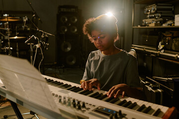 Backlight of teenage boy creating beautiful melody with professional keyboard at studio filled with...