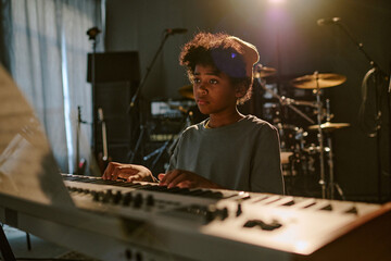 Tired teen boy looking at papers with notes while playing keyboard at dimly lit studio
