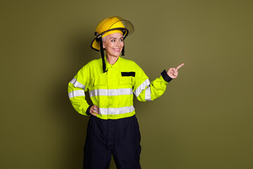 Confident woman in safety gear pointing while smiling, wearing a protective helmet and high-visibility uniform against a khaki background