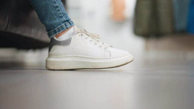 Close-up side view of lady's white canvas sneakers gently tapping right foot on the floor while seated, with blurred background showcasing modern, stylish environment