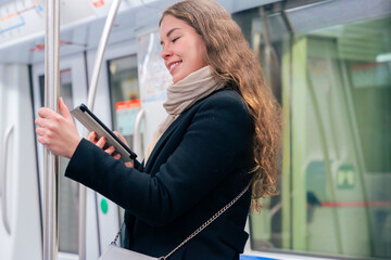  Stylish young woman in a black coat and beige scarf reading on an e-reader while standing inside a...