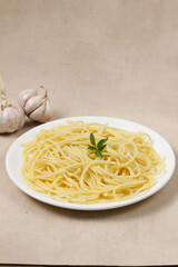 A plate of perfectly cooked spaghetti, garnished with a sprig of parsley, sits on a beige background, accompanied by garlic bulbs.
