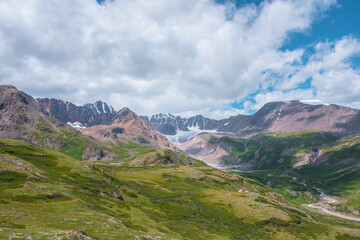 Fototapeta premium Colorful vast scenery of wide green alpine valley with panoramic view to large snow-capped top, sharp rockies, rocky pointy peak, snowy mountain range and big glacier tongue far away under cloudy sky.
