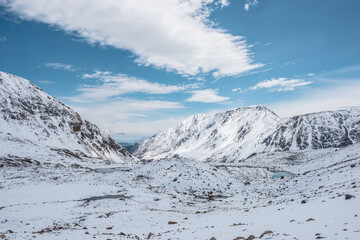 Cold snow-white landscape of snowy valley with alpine lakes against snow-covered rocky mountain range under clouds in blue sky. Glacial lakes, pure white snow and stones in high mountains in sunny day