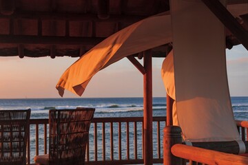 A close-up of a wooden gazebo with white curtains swaying in the sea breeze at sunset. The golden...