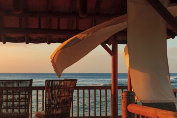 A close-up of a wooden gazebo with white curtains swaying in the sea breeze at sunset. The golden...