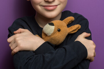 Adorable child with arms crossed, hugging a cute kangaroo plushie on a purple background. Represents themes of love, care, and the warmth of childhood memories.
