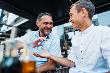 Handsome and happy father and his teenage son sitting in a restaurant and talking.