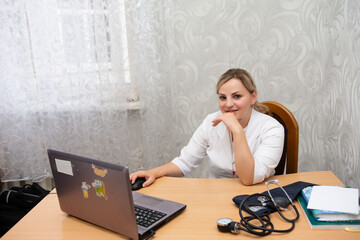A medical professional in a white lab coat is seated at a desk, working on a laptop and handling a...