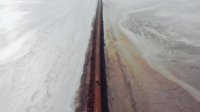 Aerial view of an isolated dirt road stretching along Konya Salt Lake. The cracked arid land and white salt layer on either side of the road reveal striking contrasts in nature.