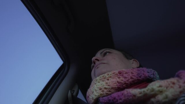 video of woman in car looking out of window, looking sad and pensive. She is wearing a colourful scarf and through the window we can see the sky.