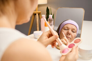 A woman focuses on her reflection while applying blush from a palette in front of a well-lit...