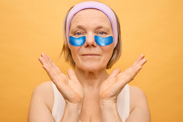 A mature woman enjoys a moment of self-care, applying blue eye patches under her eyes while showcasing a peaceful smile. The bright yellow background enhances her uplifting mood