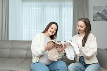 Young pregnant woman and her female friend choosing baby clothes while sitting on a couch. Concept of motherhood, friendship, preparation, excitement, and bonding.