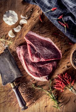 Two beef steaks on a butcher's board, surrounded by spices, herbs and garlic