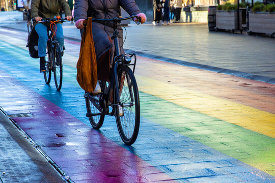 Two cyclists ride stylish black bicycles across a vibrant rainbow crosswalk on a Regenboogvlag in the Grote Marktstraat in the Hague, Nethrlands.
