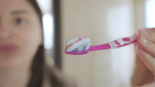 Hand holds pink toothpaste by the sink in the bathroom. Hands with toothpaste and brush, starting oral care routine.