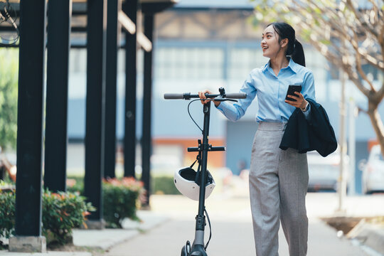 Professional businesswoman commuting, riding electric scooter, checking smartphone, wearing light blue shirt, gray pants, jacket draped over arm, helmet secured on handlebar - Powered by Adobe