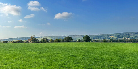 A vast green field stretches out under a blue sky, dotted with a few fluffy clouds. In the background, rolling hills and scattered trees create a serene and picturesque landscape near, Wilsden, UK