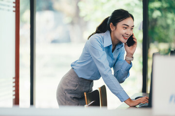 Fototapeta premium Professional Asian woman multitasking, communicating via smartphone while typing on laptop in contemporary corporate workspace