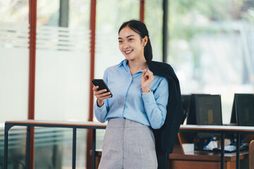 Fototapeta premium Professional Asian businesswoman holding jacket, talking on smartphone, standing confidently near contemporary office window with natural light