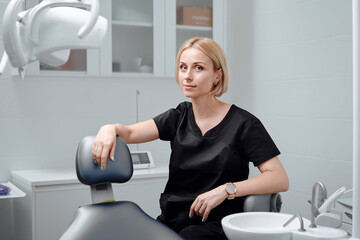 Young blonde woman dentist posing at modern dental cabinet next to dental chair. Pretty stomatologist at dental clinic