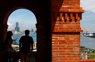 Tourists stand at the arch corridor of the historic architecture of Former British Consulate in Takao, Taiwan & enjoy a panoramic view of Kaohsiung Harbor with the landmark 85 Sky Tower in background