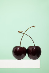 Vertical image in close up  of a pair of cherries on a white wooden shelf on a light green background, small round red fruit
