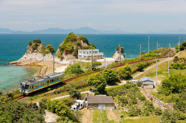 A local train travels on JR Yosan Railway  along the beautiful coastline and Komogahana (a small headland) extends into the blue water of Seto Inland Sea near Oura, in Matsuyama, Ehime, Shikoku, Japan