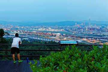 Tourist on a viewing platform enjoying a panorama of Downtown Taipei, the vibrant capital city of Taiwan, with 101 Tower amid skyscrapers in Xinyi District & a riverside park by Keelung River at dusk