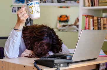 Exhausted young female employee feeling fatigue lying on table and lifting cup of coffee lack of...