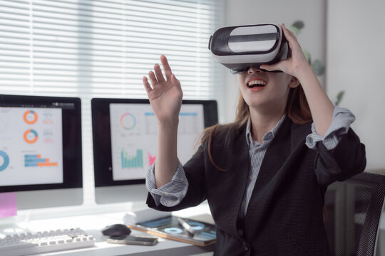 Businesswoman wearing virtual reality headset is interacting with metaverse while working with charts and graphs displayed on computer screen in office