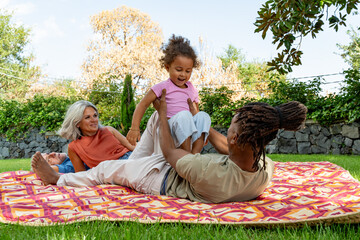 A happy multi-generational and diverse family enjoying a picnic outdoors. A father plays with his daughter while the grandparents watch, representing love, bonding and family legacy.