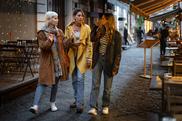 Three stylish women walking and talking in a cozy city street with festive evening lights